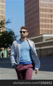 Portrait of a young bearded man with sunglasses, model of fashion, in urban background wearing casual clothes while walking with hand on pocket