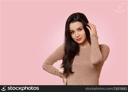 portrait of a young beautiful Asian brunette girl in a light brown jacket stands with her hand touching her hair. on a pink isolated background with copyspace.