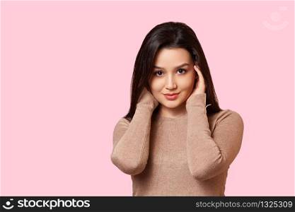 portrait of a young beautiful Asian brunette girl in a light brown jacket holding her hands over her face . on a pink isolated background with copyspace.