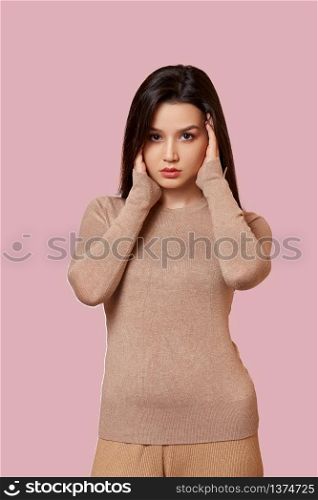 portrait of a young beautiful Asian brunette girl in a light brown jacket holding her hands over her face . on a pink isolated background with copyspace