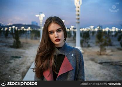 portrait of a young beautiful brunette girl in a gray coat against the sunset and night lights