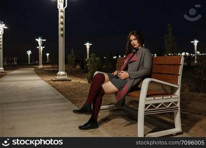 portrait of a young beautiful brunette girl in a gray coat and Burgundy stockings sitting on a bench with a vintage camera in her hands at night under the light of lanterns
