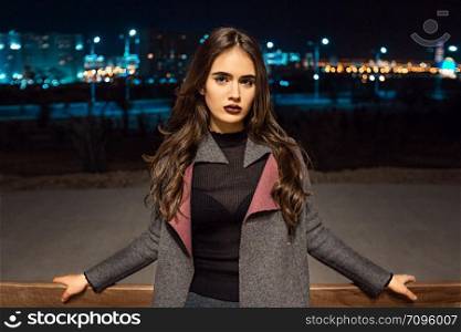 portrait of a young beautiful brunette girl in a gray coat and Burgundy stockings standing near a bench in the light of lanterns at night