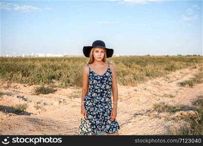 portrait of a young beautiful caucasian blonde girl in a blue dress with a floral print and a black hat standing on a sandy village road near a grape field in the summer during sunset