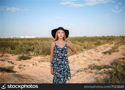 portrait of a young beautiful caucasian blonde girl in a blue dress with a floral print and a black hat standing on a sandy village road near a grape field in the summer during sunset