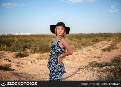 portrait of a young beautiful caucasian blonde girl in a blue dress with a floral print and a black hat standing on a sandy village road near a grape field in the summer during sunset