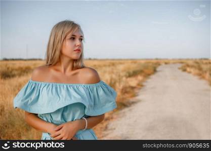 portrait of a young beautiful caucasian blonde girl in a light blue dress standing on a field with sun-dried grass next to a small country road