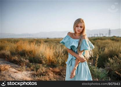portrait of a young beautiful caucasian blonde girl in a light blue dress standing on a field with sun-dried grass next to a small country road