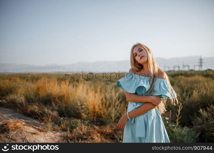 portrait of a young beautiful caucasian blonde girl in a light blue dress standing on a field with sun-dried grass next to a small country road