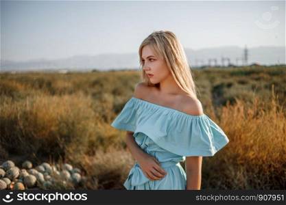 portrait of a young beautiful caucasian blonde girl in a light blue dress standing on a watermelon field with sun-dried grass during sunset