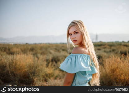 portrait of a young beautiful caucasian blonde girl in a light blue dress standing on a watermelon field with sun-dried grass during sunset