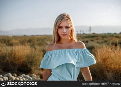 portrait of a young beautiful caucasian blonde girl in a light blue dress standing on a watermelon field with sun-dried grass during sunset