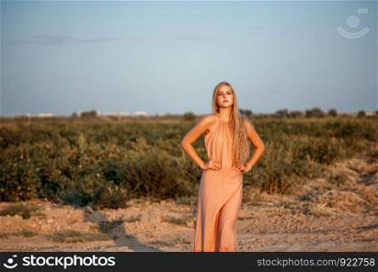 portrait of a young beautiful Caucasian blonde woman in a pink dress in a deserted field against the sunset