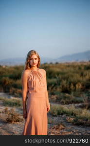 portrait of a young beautiful Caucasian blonde woman in a pink dress in a deserted field against the sunset