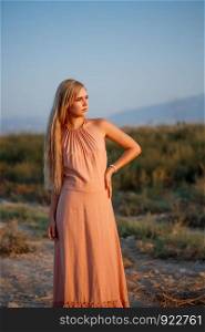 portrait of a young beautiful Caucasian blonde woman in a pink dress in a deserted field against the sunset