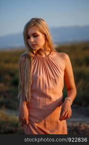 portrait of a young beautiful Caucasian blonde woman in a pink dress in a deserted field against the sunset