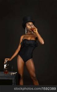 Portrait of a young black female with long dreadlocks & beautiful makeup drinking whiskey in a studio with dark background wearing silver jewelry, top hat & black bodysuit while smoking a joint.