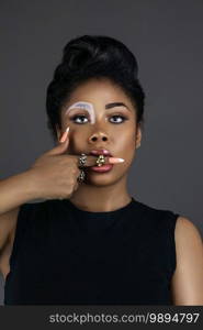 Portrait of a young black female with long dreadlocks, beautiful makeup and moist lips posing by herself in a studio with brown background wearing jewelry, denim shorts, a body top   jacket.