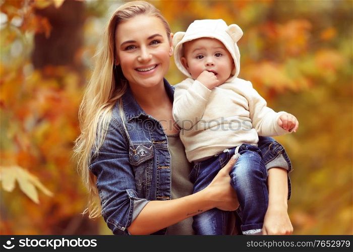 Portrait of a young blond mother with cute little son in autumn park over dry tree foliage background, happy family with pleasure spending time together