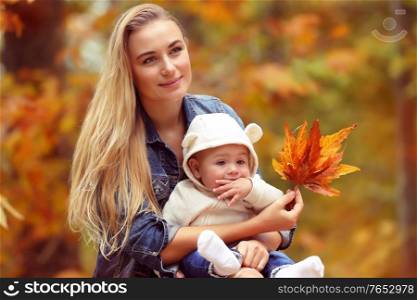 Portrait of a young blond mother with cute little son sitting in the autumn park over dry tree foliage background, happy family with pleasure spending time together