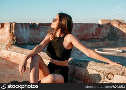 portrait of a young brunette girl in black sneakers,black shorts and a black short top sitting on the roof and enjoying the sunset