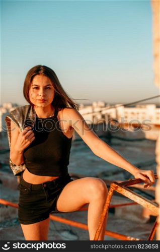 portrait of a young brunette girl in black sneakers,black shorts and a black short top standing on the roof and enjoying the sunset
