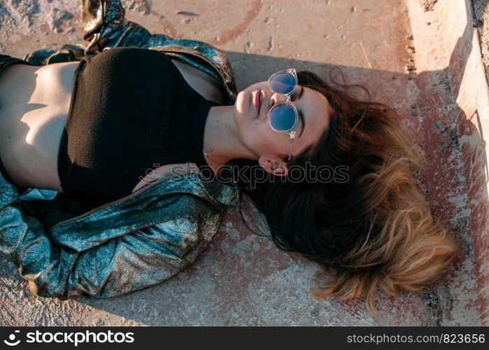 portrait of a young brunette girl with a beautiful body in black clothes and a silver jacket lying on the roof and enjoying the sunset