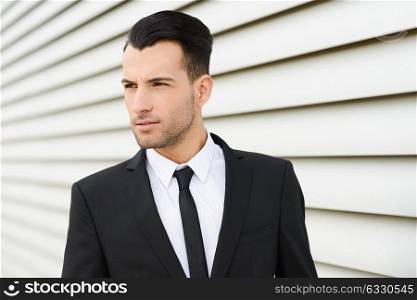 Portrait of a young businessman near a office building wearing black suit and tie with modern haircut