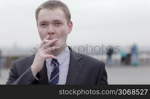 Portrait of a young Caucasian businessman wearing a formal elegant gray suit, thinking while smoking a cigarette outdoors