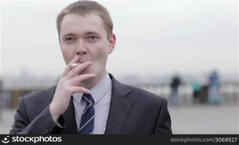 Portrait of a young Caucasian businessman wearing a formal elegant gray suit, thinking while smoking a cigarette outdoors