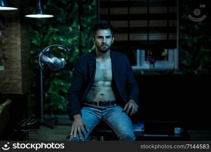 portrait of a young Caucasian guy in a black jacket on a naked body and gray jeans sitting in a stylish interior in the light of neon lights
