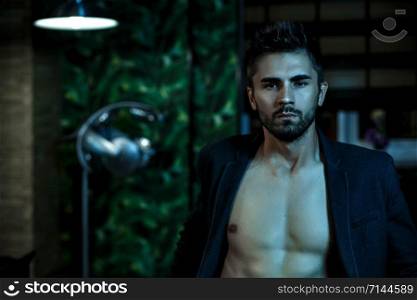 portrait of a young Caucasian guy in a black jacket on a naked body and gray jeans sitting in a stylish interior in the light of neon lights