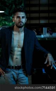 portrait of a young Caucasian guy in a black jacket on a naked body and gray jeans stands in a stylish interior in the light of neon lights