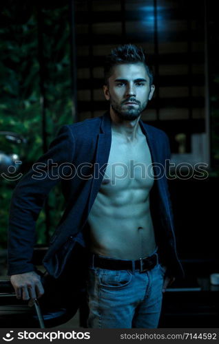 portrait of a young Caucasian guy in a black jacket on a naked body and gray jeans stands in a stylish interior in the light of neon lights