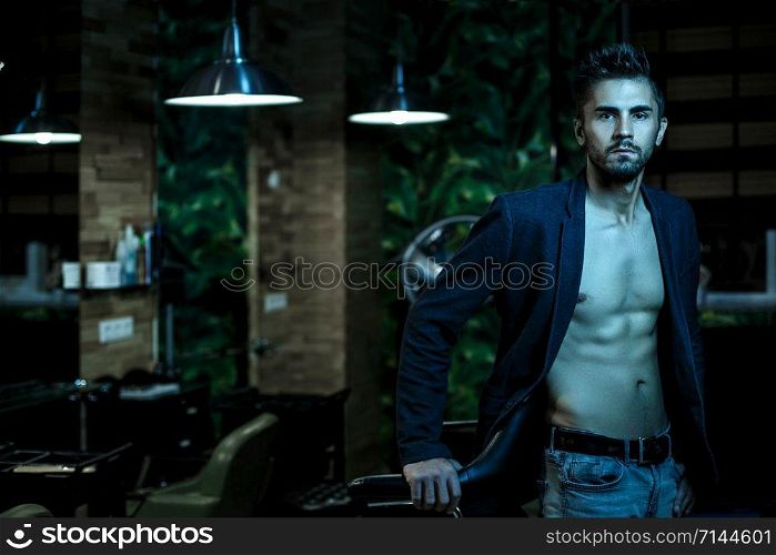 portrait of a young Caucasian guy in a black jacket on a naked body and gray jeans stands in a stylish interior in the light of neon lights