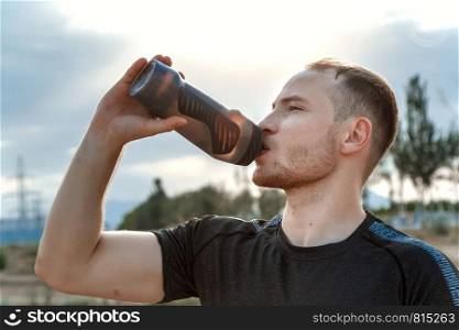 portrait of a young Caucasian guy in a black t-shirt and black shorts drinking water from a bottle after or before training