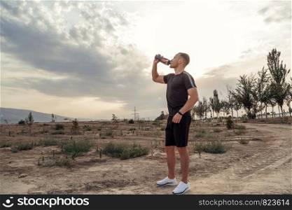 portrait of a young Caucasian guy in a black t-shirt and black shorts drinking water from a bottle after or before training