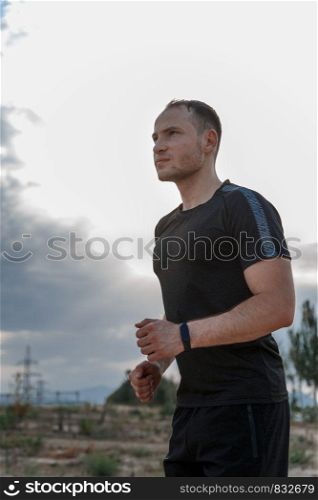 portrait of a young Caucasian guy in a black t-shirt and black shorts running over rough terrain during sunset