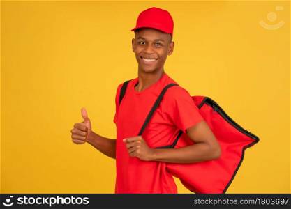 Portrait of a young delivery man looking at the camera and showing thumb up while standing against isolated yellow background. Delivery service concept.