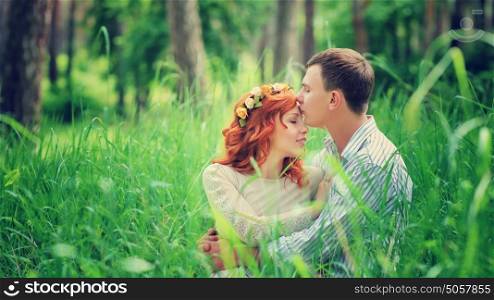Portrait of a young gentle couple sitting on a fresh green grass in the forest, kissing and enjoying spending time together
