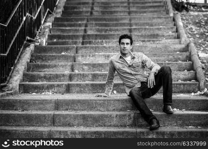Portrait of a young handsome man, model of fashion, with formal hairstyle sitting in urban stairs, wearing casual clothes. Blue eyes guy in urban background.