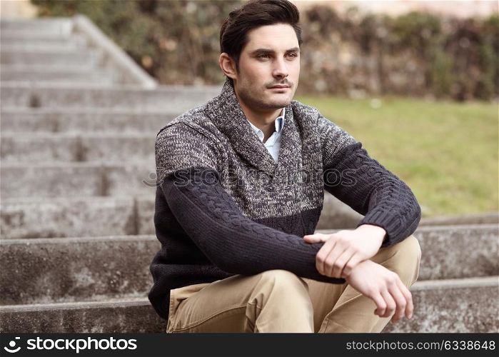 Portrait of a young handsome man, model of fashion, with modern hairstyle sitting on stairs, wearing casual clothes.