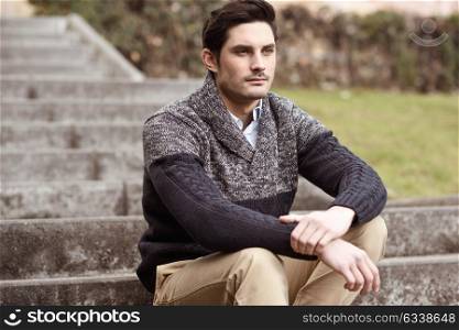 Portrait of a young handsome man, model of fashion, with modern hairstyle sitting on stairs, wearing casual clothes.