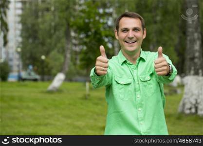 Portrait of a young handsome man with thumb up in a green shirt on the background of summer park