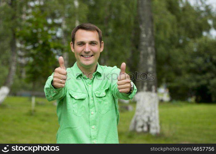 Portrait of a young handsome man with thumb up in a green shirt on the background of summer park