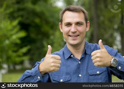 Portrait of a young handsome man with thumb up in a green shirt on the background of summer park