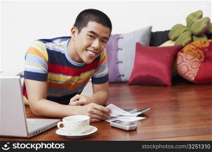 Portrait of a young man holding a bill and lying in front of a laptop