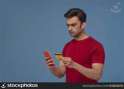 Portrait of a young man holding a credit card looking at his phone with a shocked expression.