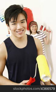 Portrait of a young man holding a paint roller with a young woman standing in the background