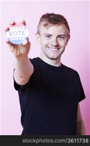 Portrait of a young man holding a VOTE badge up to the camera against pink background